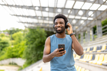 Fototapeta premium Young athletic man smiling, adjusting wireless headphones while holding a smartphone, listening to music or a podcast, standing outdoors at a sports stadium getting ready for a workout