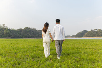 Naklejka premium A young couple of asian ethnicity walks hand in hand on a lush green grassland, facing distant hills and water in natural outdoor scenery.