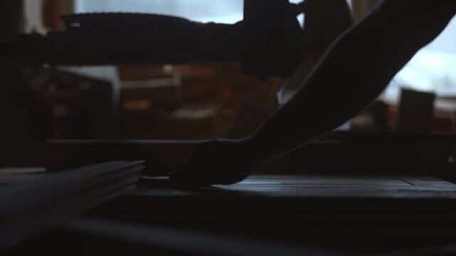 Silhouetted hand reaching toward stacked papers on low tabletop beside rail inside dim workshop with strong window backlight and muted shadows, with hand, arm, paper, tabletop detail