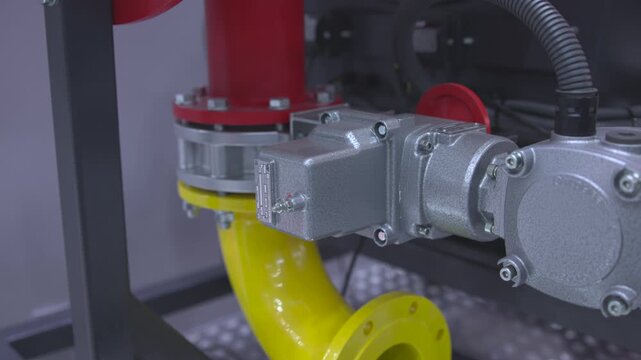 Close-up industrial gearbox mounted to flanged yellow pipe with red coupling inside utility room, oblique viewpoint showing bolts, conduit, textured cast-metal finish and rigid mechanical assembly