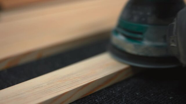 Handheld orbital sander pressing onto pine slats with circular pad, visible dust, warm workshop light, tight close-up framing, textured surface, operator glove edge, diagonal composition