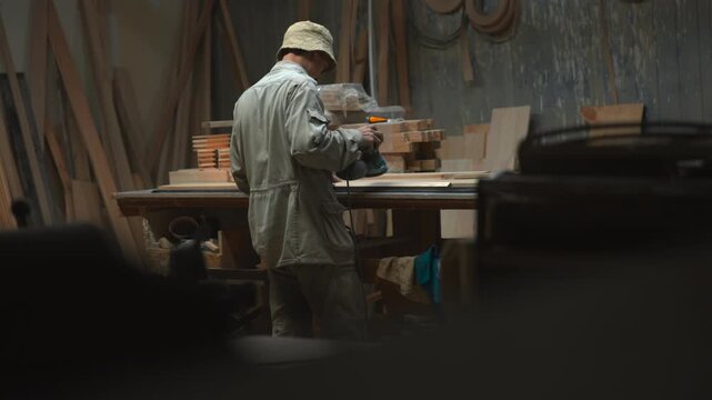 Rear view of woodworker shaping stacked lumber at workbench inside rustic workshop, using handheld power tool amid dusty boards, worn walls, muted light, and deep foreground blur