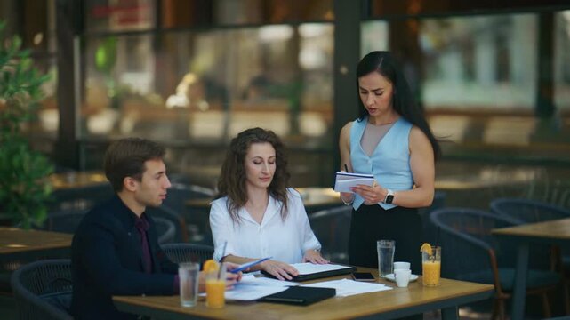 Professionals engage in an outdoor business meeting at a bustling cafe. They are seen discussing important documents and strategizing over drinks.
