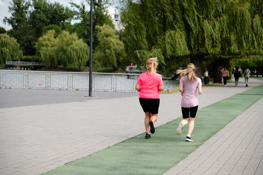 Women jogging along a path beside a park with trees and water on a sunny day