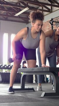 Vertical video: Bracing left knee, hand on bench, woman tank rowing dumbbell training, spotter near
