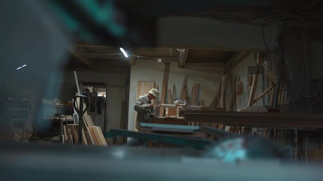 Woodworker finishing stacked boards at cluttered workbench inside busy workshop under cool fluorescent light, handheld sander moving over wood while craftsman leans in with focused posture