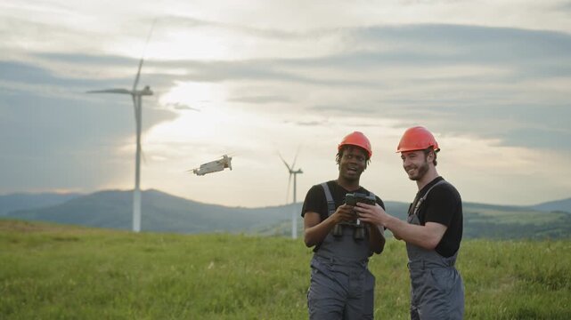 Two technicians in helmets operate a drone near wind turbines in grassy field. They monitor renewable energy systems, collect data, and ensure clean electricity through modern technology and solutions