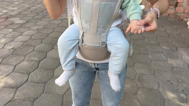 a mother holding her baby's hands while carrying them in a front-facing baby carrier outdoors. Modern parenting