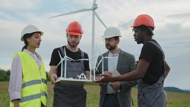 Engineers in helmets discuss wind turbine model near full-scale turbine. Their teamwork emphasizes renewable energy innovation, clean electricity, and sustainable technology for modern industry