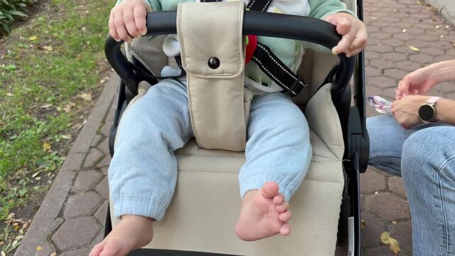 a baby sitting in a stroller kicking bare feet while mother handles wipes outdoors. Everyday parenting