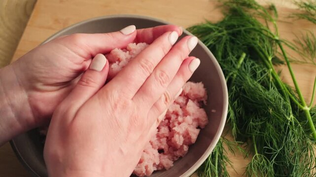 Close-up of hands combining minced meat with fresh dill in a bowl during homemade meal preparation. Traditional cooking process, natural ingredients, kitchen workspace, healthy food concept. High