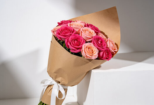 Pink peach rose bouquet wrapped brown craft paper white ribbon bow minimalist white studio setting bright light shadows