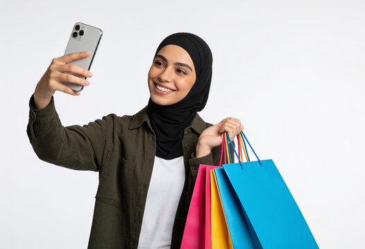 Smiling young Muslim woman in hijab taking selfie with smartphone holding colorful shopping bags studio portrait