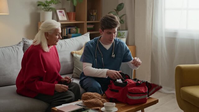 Young Caucasian male nurse opening medical bag and showing blood pressure monitor to senior Caucasian woman at home. Ideal for promoting home healthcare services and geriatric care