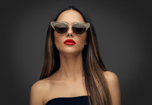 A stylish studio portrait of a glamorous woman with long, straight brown hair wearing chic cat-eye sunglasses and bold red lipstick