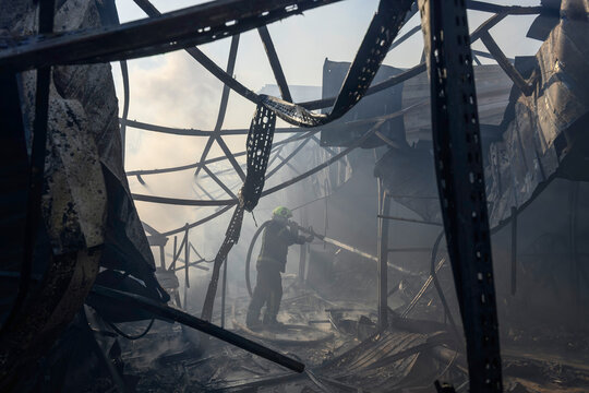 Firefighter in protective gear extinguishes a structural fire with a high-pressure fire hose amid dense smoke, flames, twisted metal debris, collapsed structures, and hazardous conditions.