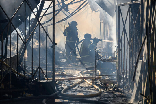 Firefighter in protective gear extinguishes a structural fire with a high-pressure fire hose amid dense smoke, flames, twisted metal debris, collapsed structures, and hazardous conditions.