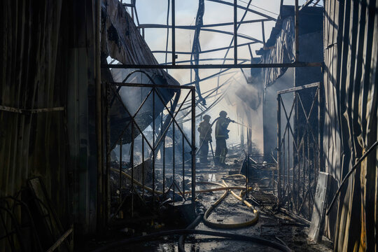 Firefighter in protective gear extinguishes a structural fire with a high-pressure fire hose amid dense smoke, flames, twisted metal debris, collapsed structures, and hazardous conditions.