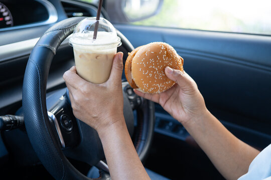 Asian lady holding hamburger and ice coffee to eat in car, dangerous and risk an accident.
