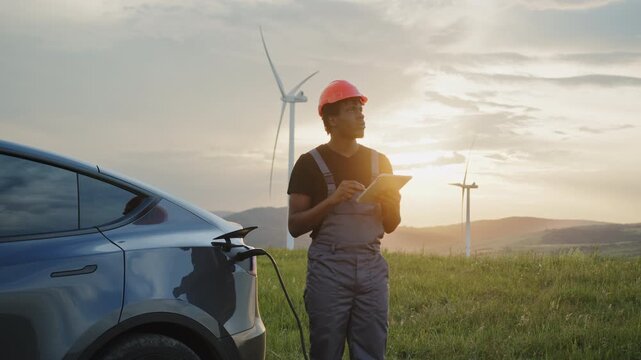 Man in helmet and overalls checks tablet near electric car charging at wind farm. The scene symbolizes renewable energy, clean electricity, and sustainable technology driving future mobility.