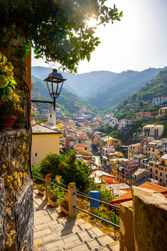Riomaggiore Village is seen from a high point in the Cinque Terre region of Italy. The scene shows houses stacked on the hillside with mountains in the background.