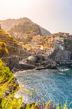 Houses sit on the hillside of Manarola Village. The village overlooks the sea. Bright colors shine in the sunlight. Waves crash against the rocks below in Cinque Terre, Italy.