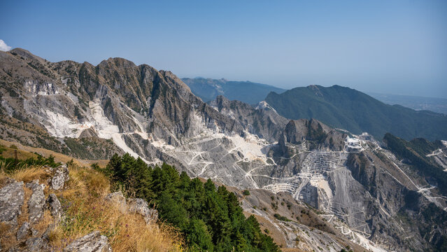Workers extract white Carrara marble from quarries in Italy. The scene captures the steps and paths carved into steep mountains. The sky is clear and the land is rugged.