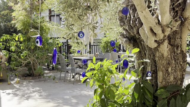 Traditional Turkish blue glass evil eye amulets Nazar Boncugu hanging on olive tree branches swaying in the wind at a cozy outdoor cafe garden