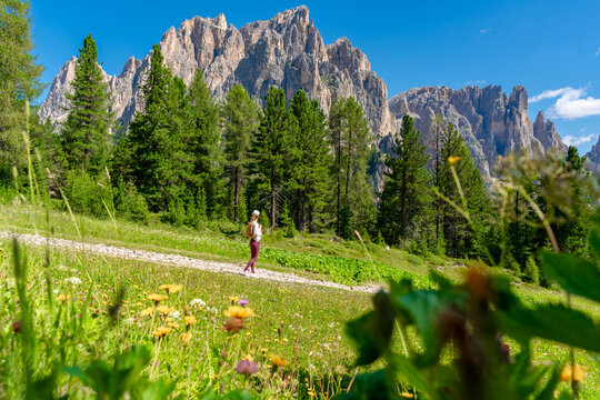 Young woman with a backpack walking along a mountain trail across a green alpine meadow bordered by pine trees. The image conveys active lifestyle, hiking, eco tourism, healthy recreation