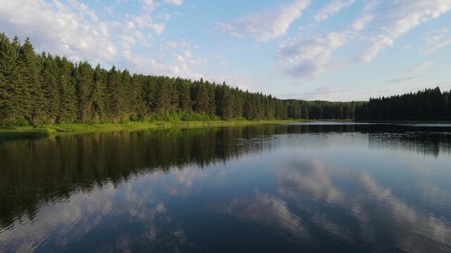 Drone flying low above a calm northern park lake with summer clouds reflecting in the water. Smal waves distort the reflection.
