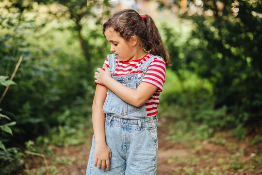 Mosquitoes are biting a girl, and she is scratching her arm in a summer forest. The child, wearing denim overalls and a striped T-shirt, is standing on a forest path.