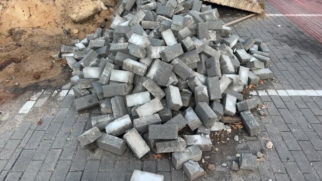 Pile of grey concrete paving stones stacked on a construction site near a dirt patch and paved walkway. Roadwork, landscaping, building materials