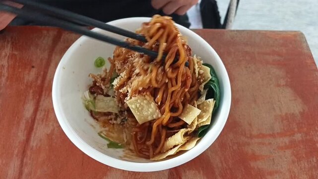 Footage of spicy fried noodles in a white bowl being lifted with chopsticks on a wooden table.
