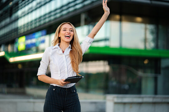 Happy businesswoman waving hand while standing on city street, holding digital tablet