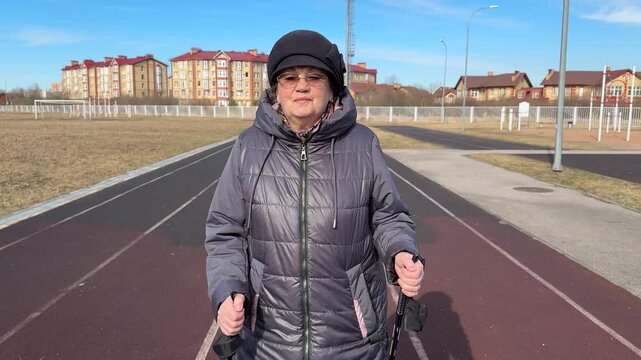 Senior woman Nordic walking outdoors treadmill, brisk spring morning wearing puffer jacket beanie and glasses using poles on track
