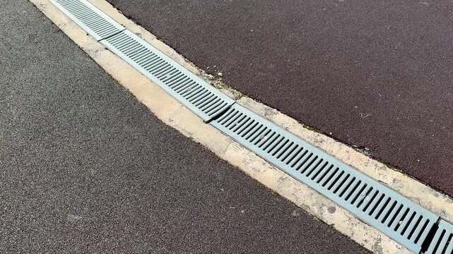 Galvanized drainage grate on asphalt, linear perforated metal slats running along track joint at stadium street closeup top down