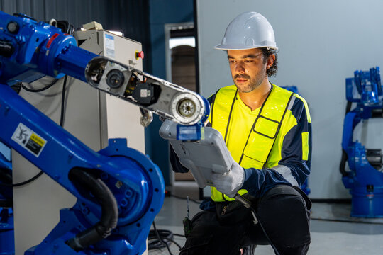 Engineer in yellow safety vest uses tablet to program blue robotic arm in industrial workshop with corrugated metal walls