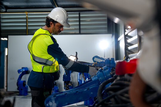 Engineer in yellow vest and white helmet adjusts blue robotic arm with focused intensity in clean industrial workshop