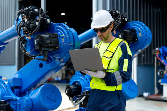 Engineer in yellow vest and white helmet programs blue robotic arm with laptop in industrial workshop with corrugated metal walls