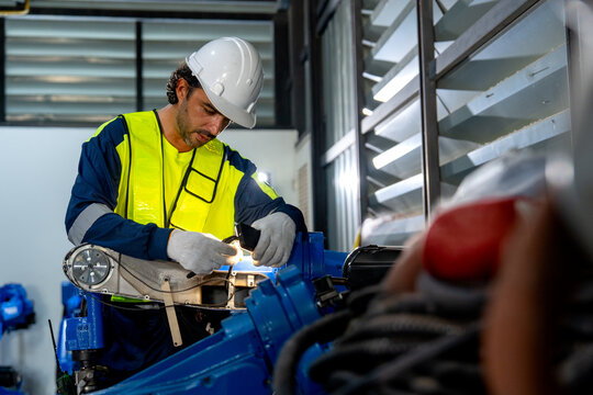 Engineer in yellow vest and white helmet carefully adjusts blue robotic arm in industrial workshop with corrugated metal walls