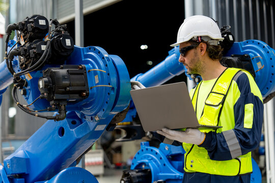 Engineer in yellow vest and white helmet programs blue robotic arm using laptop in industrial workshop with corrugated metal walls