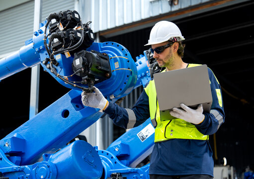 Engineer in safety gear calibrates blue robotic arm with laptop under industrial shed lighting