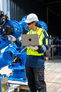 Engineer in safety gear calibrates blue robotic arm with laptop under industrial shed lighting
