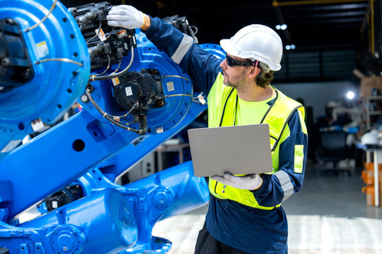 Engineer in safety gear adjusts blue robotic arm while holding laptop in industrial workshop with corrugated walls