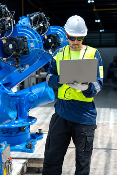 Engineer in safety gear reviews laptop data beside towering blue industrial machinery under bright workshop lighting