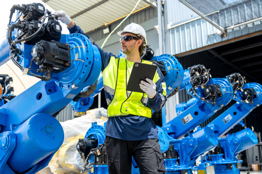 Engineer in safety gear inspects blue robotic arm with tablet under bright industrial shed lighting