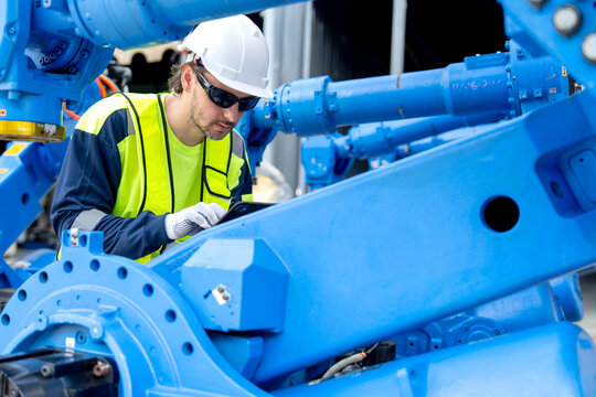 Engineer in safety gear examines blue robotic arm with tablet under industrial shed lighting