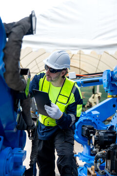 Engineer in safety gear checks tablet beside blue robotic arm under industrial tent lighting