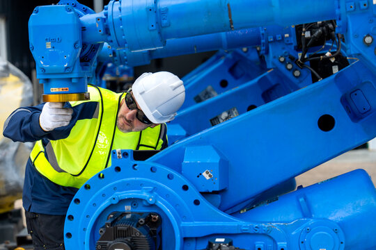 Engineer in safety gear adjusts blue robotic arm while holding laptop in industrial workshop with corrugated walls