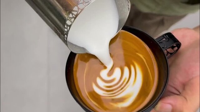 Barista pouring steamed milk from metal pitcher to create detailed swan latte art pattern in dark cup